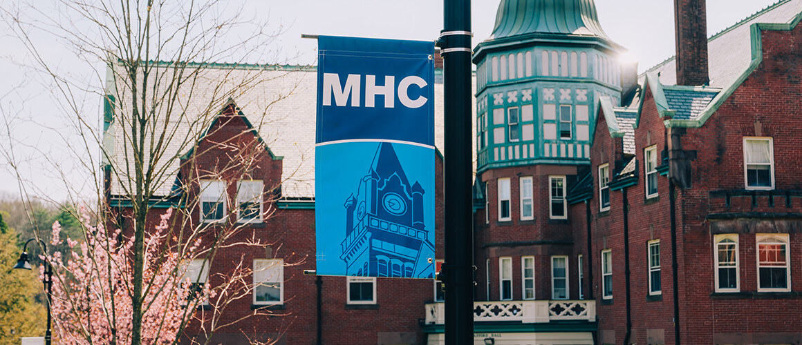 Banner on a light pole in front of Safford Hall with a newly blooming tree in the background. Mount Holyoke campus, April 2025. Photo by Max Wilhelm.