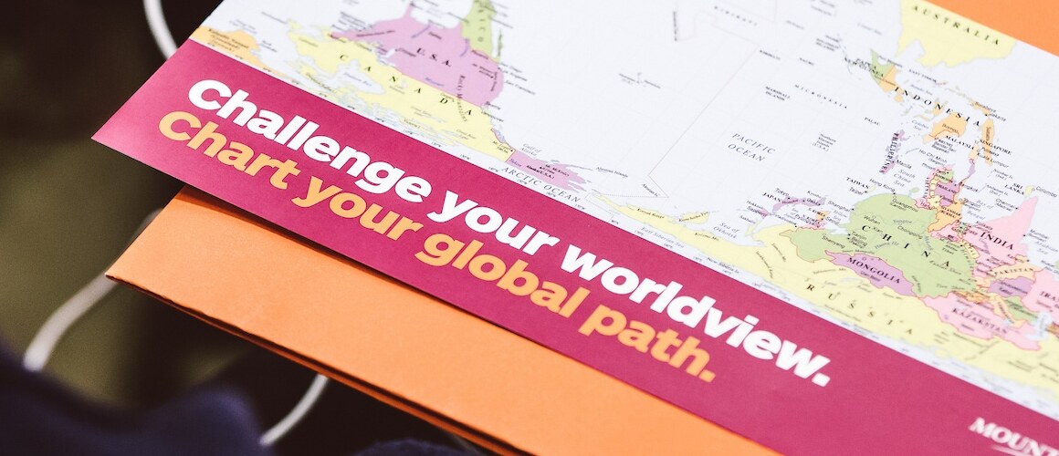 A look over a student's shoulder, laptop and flyer in view, it reads: Challenge your worldview. Chart your global path.