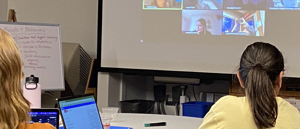 Several students listen to a lecture in a classroom. A teacher stands at a podium, and a screen shows faces of online students from across the state.