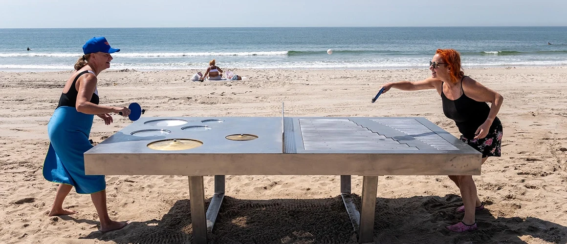 Two women volley a ball on a stainless steel Ping-Pong table embedded with tom drums, cymbals, a handpan and the bars of a glockenspiel.