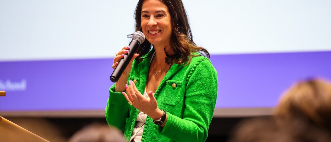 Caroline Nobo ’07, wearing a bright green jacket and holding a microphone, speaks in front of an audience.
