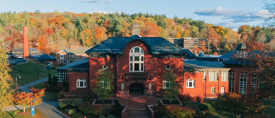 An aerial view of Mount Holyoke’s Blanchard Hall and Community Center in waning afternoon light, with colorful fall foliage and a blue sky in the background.